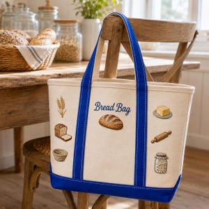 May include: A cream-colored canvas tote bag with royal blue straps and base. The front features the text "Bread Bag" in blue script, surrounded by embroidered illustrations of wheat, a loaf of bread, a basket, butter, a rolling pin, and a jar.
