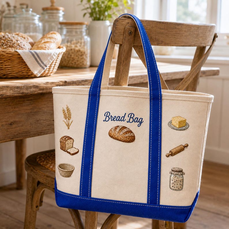 May include: A cream-colored canvas tote bag with royal blue straps and base. The front features the text "Bread Bag" in blue script, surrounded by embroidered illustrations of wheat, a loaf of bread, a basket, butter, a rolling pin, and a jar.