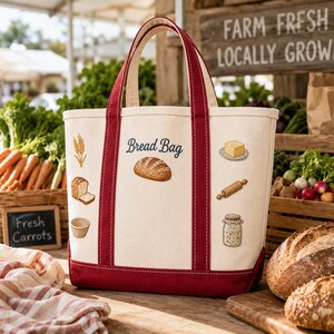 May include: A cream-colored canvas tote bag with dark red trim and handles. The bag features the embroidered text "Bread Bag" above a loaf of bread, surrounded by small embroidered icons of wheat, sliced bread, a basket, butter, a rolling pin, and a jar.