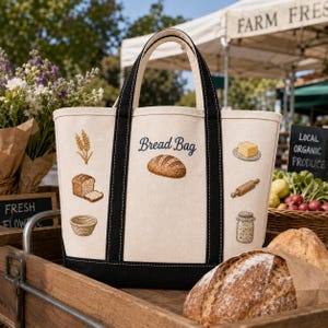 May include: A cream-colored canvas tote bag with black handles and base. The front features the text "Bread Bag" above an embroidered loaf of bread, surrounded by illustrations of wheat, sliced bread, a basket, butter, a rolling pin, and a jar.