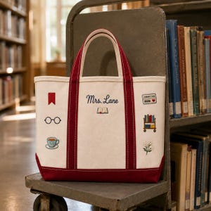 May include: A cream-colored canvas tote bag with dark red handles and a matching red base. The bag features embroidered designs including a bookmark, glasses, a teacup, a library card, a book cart, and a small flower. The text "Mrs. Lane" is embroidered in dark blue above a small open book icon.