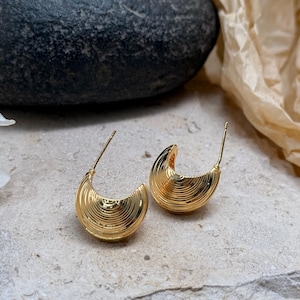 May include: A pair of gold-colored hoop earrings. Each earring has a crescent shape with a ridged, textured surface. The earrings are displayed on a light-colored, textured surface.