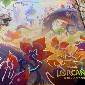 May include: A Disney Lorcana Trading Card Game playmat featuring a vibrant fantasy landscape with a character swinging on a rope. The mat has a purple border and the game's logo. The artwork includes orange and purple flowers.