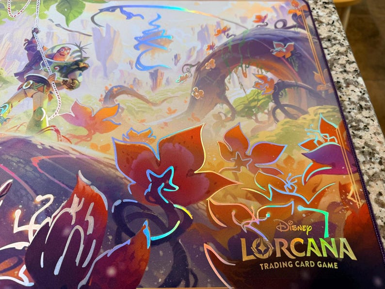 May include: A Disney Lorcana Trading Card Game playmat featuring a vibrant fantasy landscape with a character swinging on a rope. The mat has a purple border and the game's logo. The artwork includes orange and purple flowers.