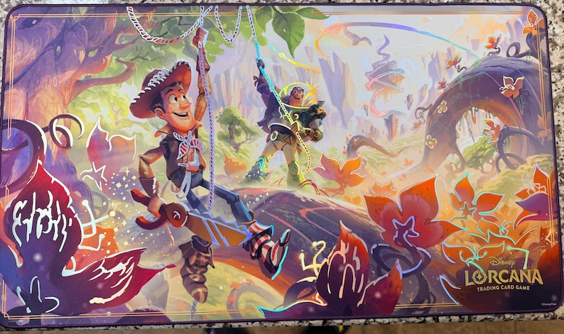 May include: A rectangular playmat featuring a vibrant illustration of characters from the Disney Lorcana Trading Card Game. The artwork depicts Woody and Buzz Lightyear in a colorful forest setting, with the game's logo in the lower right corner.
