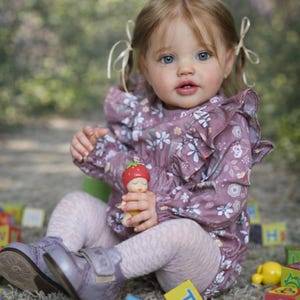 Puede incluir: Una muñeca con ojos azules y cabello rubio, vestida con un vestido con estampado floral y sosteniendo un pequeño juguete. La muñeca está sentada en una superficie con bloques de colores esparcidos.