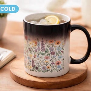 Könnte beinhalten: Eine farbwechselnde Tasse mit floralem Design. Die Tasse wechselt von Schwarz zu einem bunten Wildblumenmuster, wenn sie kalt ist. Sie enthält Eis und eine Zitronenscheibe. Die Tasse steht auf einem Holzuntersetzer.