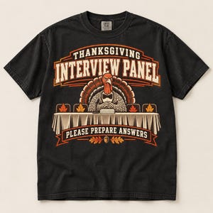 Op de afbeelding: Een zwart t-shirt met korte mouwen en een afbeelding van een kalkoen met een vlinderdas aan een tafel met een microfoon. De tekst luidt THANKSGIVING INTERVIEW PANEL en PLEASE PREPARE ANSWERS, met oranje esdoornbladeren en een eikel.