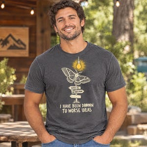 Könnte beinhalten: Ein anthrazitfarbenes Kurzarm-T-Shirt mit einer Grafik eines Nachtfalters, der auf eine leuchtende Glühbirne über einem hölzernen Wegweiser zufliegt. Der Wegweiser zeigt REPLY ALL und ONE MORE EPISODE. Darunter steht I HAVE BEEN DRAWN TO WORSE IDEAS.