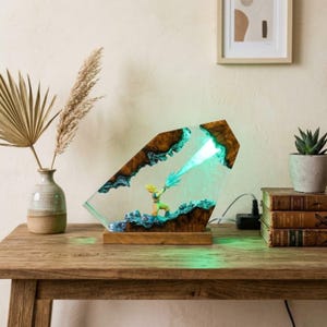 Könnte beinhalten: Eine einzigartige Harzlampe mit einer Charakterfigur, die einen blauen Energiestrahl aussendet. Die Lampe hat einen Holzsockel und einen klaren, abgewinkelten Harzkörper mit braunen und blauen Akzenten.