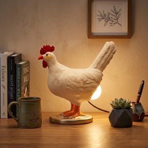 Könnte beinhalten: Eine weiße Tischlampe in Form eines Huhns mit rotem Kamm und orangefarbenen Füßen. Die Lampe hat einen runden Sockel und eine warme Glühbirne. Sie steht auf einer Holzoberfläche, neben einer Tasse, Büchern und einer kleinen Sukkulente.