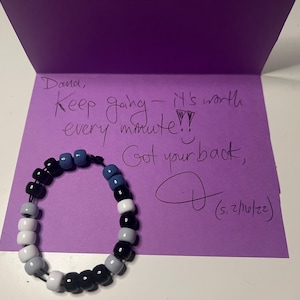 May include: A beaded bracelet with black, white, gray, and blue beads rests in front of a purple card. The card has handwritten text that reads "Keep going - it's worth every minute!!" and "Got your back,".