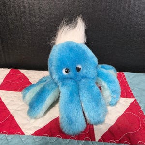 May include: A plush blue octopus toy with a white tuft of hair on its head. The octopus has black button eyes and blue and light blue striped tentacles. It is sitting on a red, white, and blue patterned quilt.