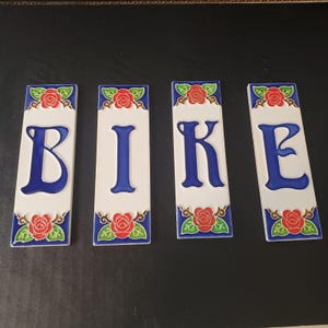 May include: Four rectangular ceramic tiles spelling "BIKE". Each tile has a white background with a blue border and the letters in blue. The top and bottom of each tile feature a red rose design.