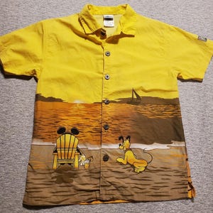 Może przedstawiać: Żółta koszula zapinana na guziki z motywem plażowym. Koszula przedstawia Myszka Miki relaksującego się na leżaku i Pluto na piasku. Na wodzie widoczny jest żaglówka. Koszula ma krótkie rękawy i kołnierzyk.