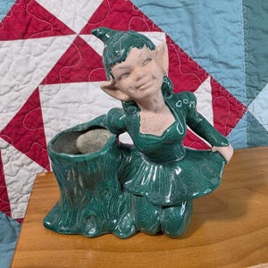 May include: A ceramic planter shaped like a forest elf with pointed ears and a hat, wearing a dark green dress. The elf is kneeling beside a hollow tree stump container with a textured bark finish and a glossy, deep green glazed surface.