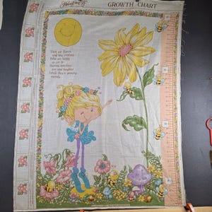 May include: A vintage fabric growth chart featuring a smiling sun, a fairy, flowers, bees, and a large sunflower. The chart includes a measurement scale in inches and the text "GROWTH CHART". The design is in pastel colors.
