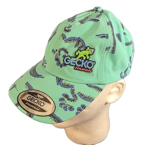 Puede incluir: Una gorra de béisbol verde claro con un patrón abstracto repetitivo en azul oscuro y marrón. El frente muestra un logotipo de Gecko Hawaii en relieve con un gecko verde brillante. Una pegatina circular en la visera dice Gecko Hawaii Est. 1979 Wear With Aloha.