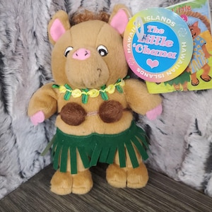 Op de afbeelding: Een pluchen varkentje met een groene graskokerrok, een bruine kokosnoot-bh en een gele en groene lei. Het varkentje heeft roze oren, snuit en voeten. Een label leest "The Little Ohana" met Hawaïaanse eilanden tekst.