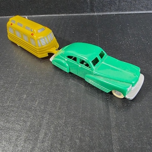 May include: A vintage toy car in green is connected to a yellow toy bus. The car has a silver bumper and white tires. The bus has a series of windows and red lights on top. The toys are on a black surface.