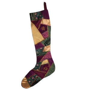 Könnte beinhalten: Ein Weihnachtsstrumpf mit einem Patchwork-Design aus violetten, dunkelgrünen und goldenen Stoffbahnen. Die Abschnitte sind durch dekorative gelbe Zickzacknähte verbunden, wobei einige Stoffbahnen kleine Blumenmuster und Stickereien aufweisen.
