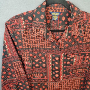 May include: A long-sleeve button-up shirt featuring a black and red patchwork pattern with geometric, floral, and paisley motifs. The shirt includes a pointed collar, a front button placket, and two chest pockets with flaps. A blue label is visible.