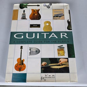 Könnte beinhalten: Ein gebundenes Buch mit dem Titel Guitar: A Complete Guide for the Player. Das weiße Cover zeigt ein Rasterlayout mit verschiedenen gitarrenbezogenen Bildern, darunter Instrumententeile, ein Marshall-Verstärker und eine Person, die eine Akustikgitarre spielt.