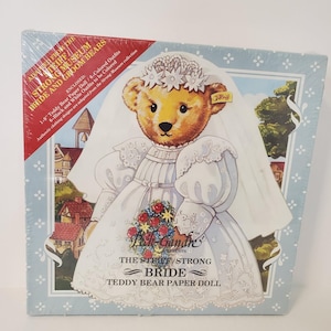 May include: A Steiff Teddy Bear paper doll in a white wedding dress with a veil and a bouquet of red and white flowers. The box reads "The Steiff/Strong Bride" and "Teddy Bear Paper Doll". The box is wrapped in plastic.
