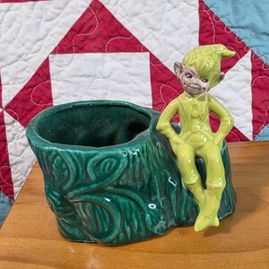 May include: A glossy dark green ceramic planter shaped like a textured tree stump with a light green elf figure sitting on the edge. The elf has a pointed hat, pointed ears, and a smiling expression, with its hands resting on its hips.