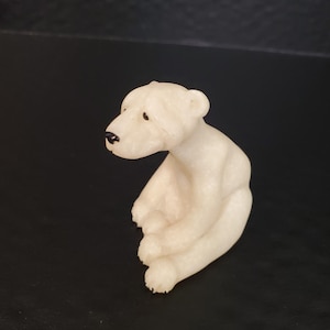 May include: A small, off-white polar bear figurine in a seated position. The bear has black eyes and a black nose. The figurine is made of a smooth material and has a detailed, realistic appearance.
