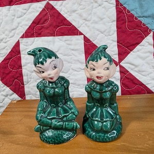 May include: A pair of vintage ceramic salt and pepper shakers shaped like seated elves. Each figure features a glossy dark green outfit with a ruffled collar, pointed hat, and painted facial details including arched eyebrows and red lips.