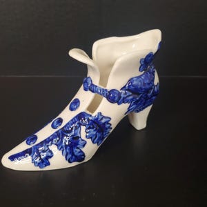 May include: A white ceramic shoe-shaped figurine featuring a glossy finish and hand-painted cobalt blue floral and leaf patterns. The design includes a pointed toe, a small heel, and a decorative strap across the top with blue circular accents.