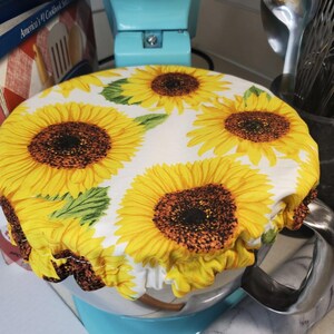 May include: A white fabric bowl cover featuring a vibrant yellow sunflower design. The cover, elasticated at the edge, fits over a light blue stand mixer bowl. The sunflowers have brown centres and green leaves.