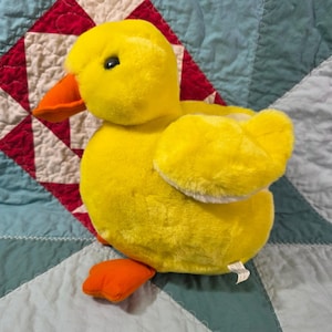 May include: A plush yellow duck toy with soft fur, featuring a bright orange beak and matching orange webbed feet. The duck has a rounded body, small wings, and dark, round eyes. A small white fabric tag is visible near the base of the toy.