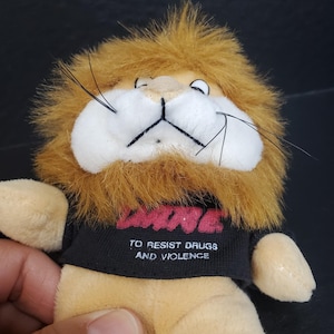 May include: A plush lion toy with a tan body, a brown mane, and long black whiskers. The lion wears a black t-shirt featuring the red text D.A.R.E. and the white text TO RESIST DRUGS AND VIOLENCE printed on the front of the fabric.