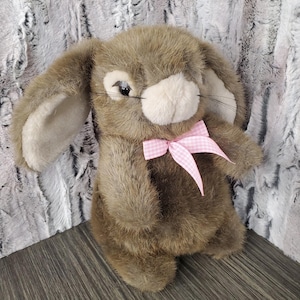 May include: A plush brown bunny with long ears and a white muzzle. The bunny has a pink and white gingham bow tied around its neck. The fur is soft and textured, giving the bunny a cuddly appearance. The bunny is a stuffed animal.