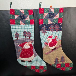 Puede incluir: Dos calcetines navideños con diseños de patchwork. Uno presenta a Papá Noel con un saco, el otro muestra a Papá Noel esquiando. Los calcetines tienen detalles en verde, rojo y azul, con patrones de árboles y estrellas.