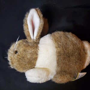 May include: A plush toy rabbit with brown and white fur. The rabbit has long ears with pink insides, a white belly, and a fluffy tail. The toy is lying on a black surface.