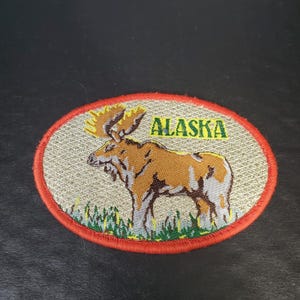 May include: An oval embroidered patch featuring a brown moose standing in green grass against a textured beige background. The word ALASKA is stitched in green letters above the moose. The patch has a bright red stitched border around the edge.