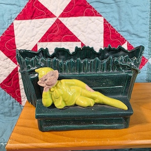 May include: A dark green ceramic planter featuring a textured fence-like design and a glossy finish. A light green, reclining elf figure with a tan face and pointed hat is attached to the front, resting on a horizontal ledge of the planter.