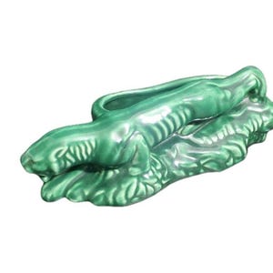 May include: A green ceramic alligator planter. The alligator is lying down with its mouth closed and has a long, open trough on its back for planting. The planter has a glossy finish and is a decorative item.