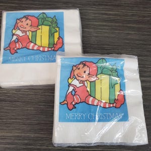 Könnte beinhalten: Zwei Packungen weiße Papierservietten. Jede Serviette hat ein blaues Quadrat mit einem Cartoon-Elf, einem gelb-grünen Geschenk und den Worten "MERRY CHRISTMAS" in weißem Text.