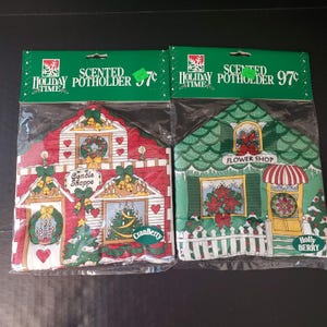 May include: Two scented holiday time potholders in plastic packaging. One is red with a candle shop design, the other is green with a flower shop design. Both have Christmas tree and holly berry accents.