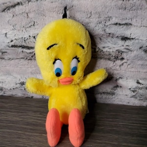 Può includere: Un peluche giallo di Titti con occhi blu, becco arancione e piedi arancioni. Il personaggio dei cartoni animati ha ciglia nere e un piccolo ciuffo di capelli neri. Il giocattolo è appoggiato su una superficie di legno.