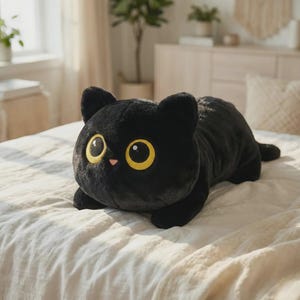 May include: A plush black cat toy with large yellow eyes and a pink nose, lying on a white bedspread. The cat has a rounded body and is in a relaxed pose. The toy is made of soft material.