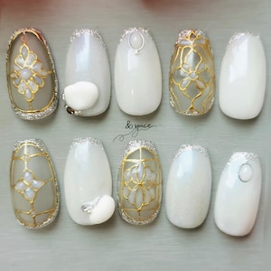 May include: A set of ten artificial nails with various designs. Some nails feature gold floral patterns, while others have white or silver glitter accents. The nails are oval-shaped and have a glossy finish. The text "& spice" is visible.