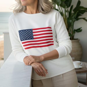 Puede incluir: Sudadera blanca de manga larga con la bandera estadounidense. La bandera presenta rayas rojas y blancas y un campo azul con estrellas blancas. La sudadera se combina con pantalones beige.