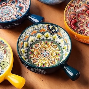 以下が含まれることがあります： 青、黄色、オレンジ、緑の色合いで、複雑な花柄と幾何学模様が特徴の、取っ手付きのカラフルなセラミックボウルのコレクション。木製の表面に配置され、装飾的なデザインが示されています。