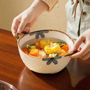 以下が含まれることがあります： 花柄と茶色の取っ手が付いたクリーム色のセラミックボウル。野菜スープが入っています。スープにはトウモロコシ、ブロッコリー、ニンジンが含まれています。ボウルは両手で持たれています。