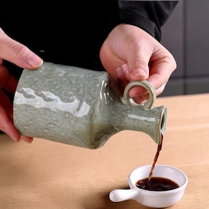 Puede incluir: Una jarra de cerámica verde oliva con asa vierte un líquido oscuro en un cuenco blanco pequeño. La jarra tiene una textura moteada y un pico curvo. El cuenco tiene un asa.